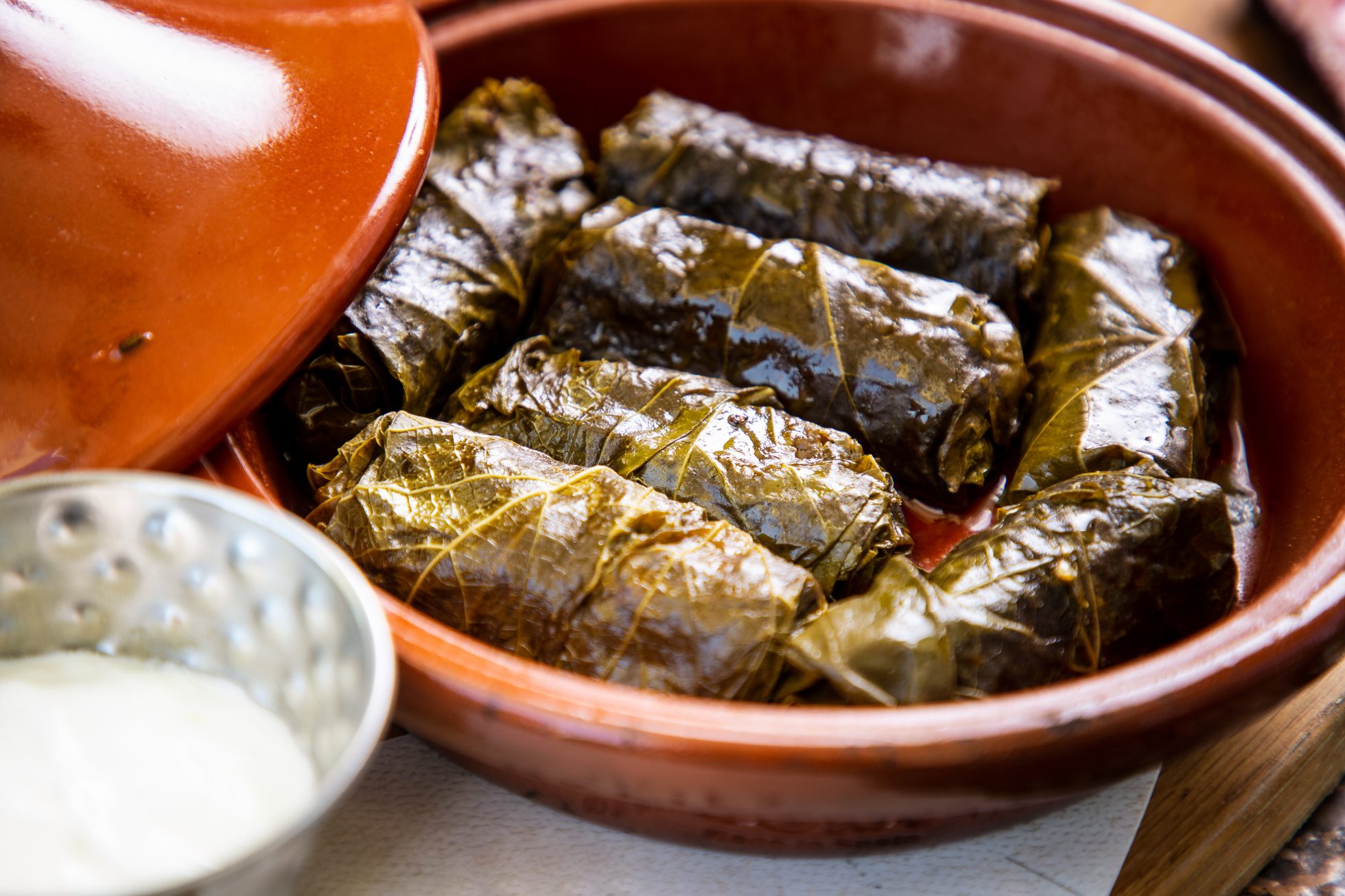Dolma w zielonych liściach winogron podawana w glinianym naczyniu, śmietana w miseczce jako sos.