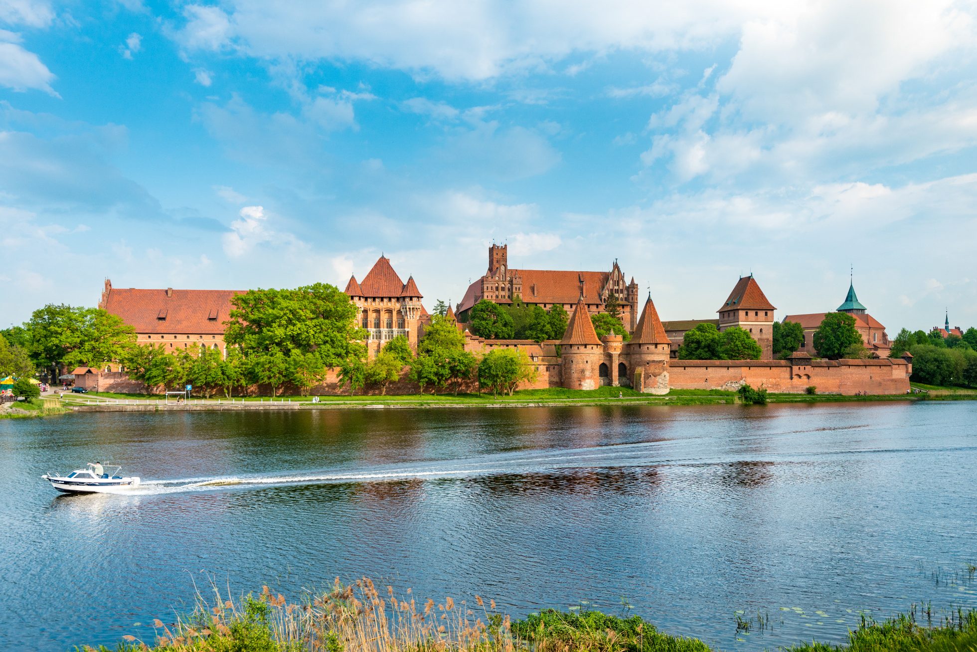 Zamek krzyżacki w Malborku (Marienburg) na Pomorzu, Polska, Europa. Obiekt wpisany na listę światowego dziedzictwa UNESCO. Błękitne niebo z chmurami w tle i rzeka Nogat na pierwszym planie.