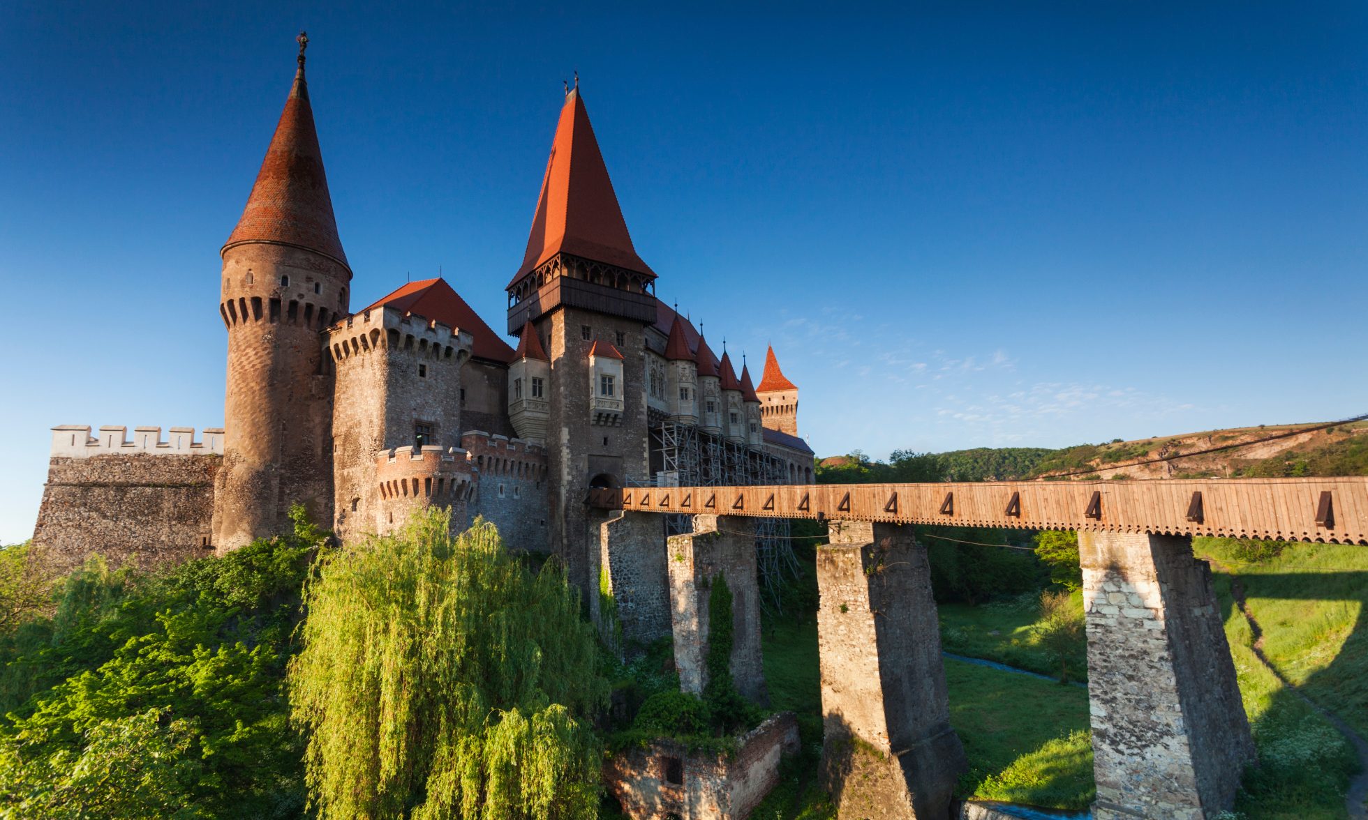 Zdjęcie przedstawia zamek Corvin w Transylwanii, Rumunia. Zamek jest otoczony górami i drzewami.