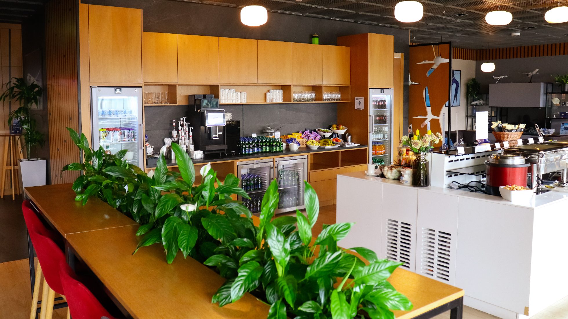 LOT Business Lounge Mazurek - wysoki, prostokątny stół z krzesłami, bar oraz wyspa z przekąskami i bemarami.