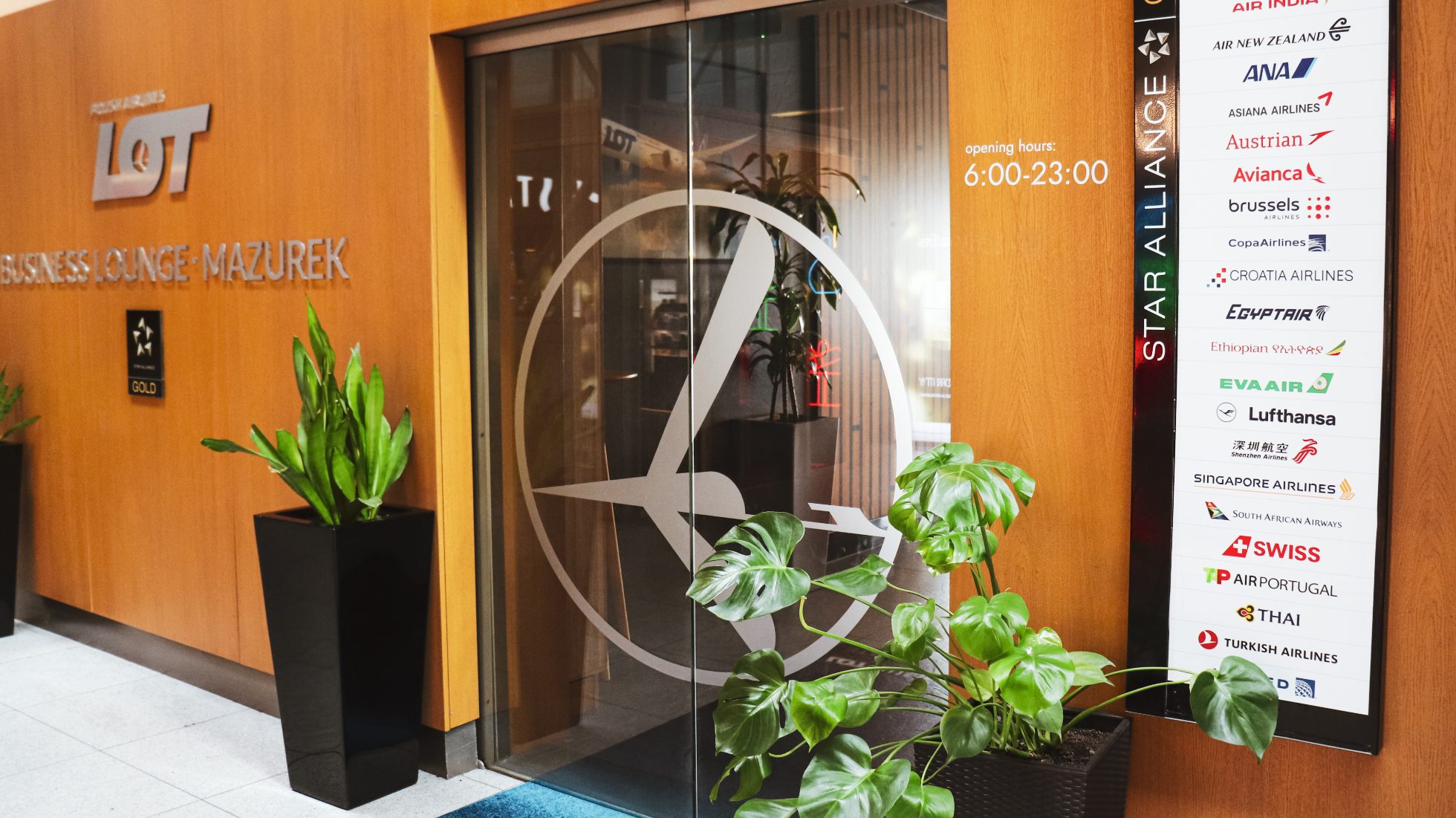 Wejście do LOT Business Lounge Mazurek - szklane drzwi z logo żurawia, tablica z liniami lotniczymi członkowskimi Star Alliance, zielony kwiat w doniczce oraz srebrny napis LOT Business Lounge Mazurek na drewnianej ścianie.