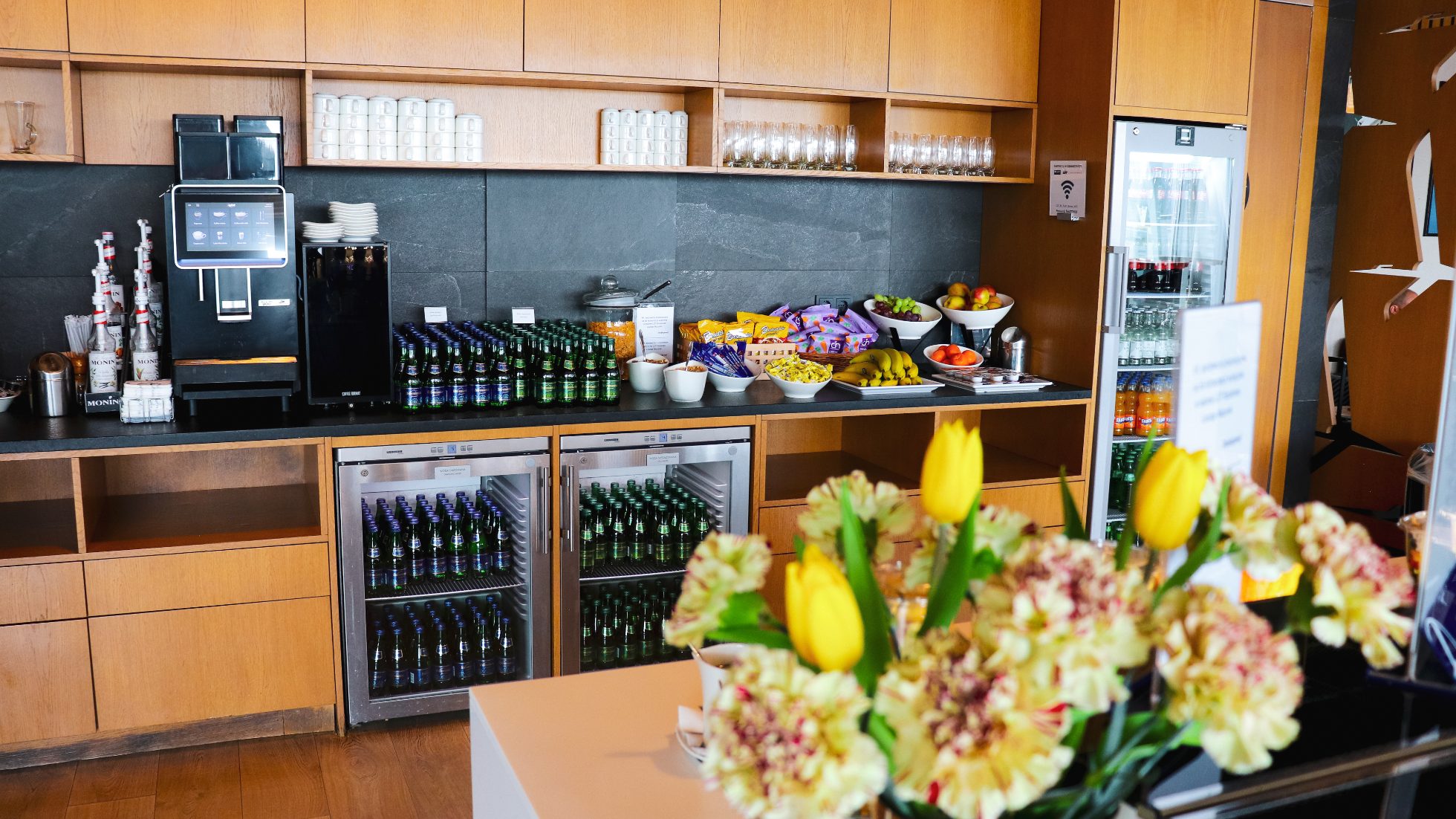 Lounge Biznesowy Mazurek - kwiaty w wazonie i bar w tle z przekąskami oraz wodą w szklanych butelkach.