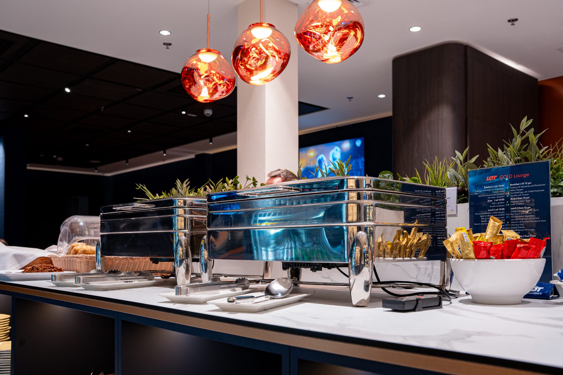Polonez Gold Lounge - otwarta przestrzeń z pomarańczowymi fotelami i kanapami, ściany w kolorze granatu z logiem żurawia. Strefa gastronomiczna z posiłkami na ciepło, przekąskami oraz napojami. Strefa wypoczynku z fotelami i przygaszonym oświetleniem 
