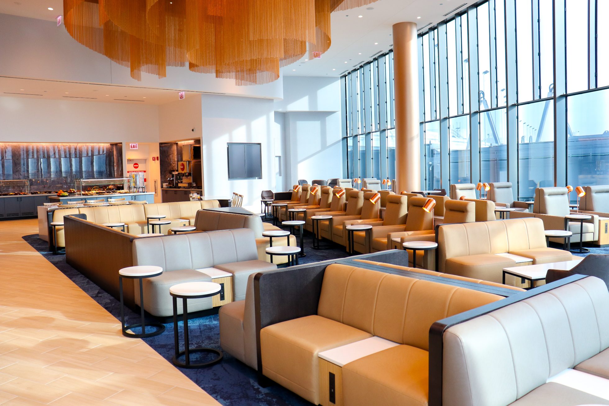 LOT Business Lounge w Chicago - recepcja z podświetlanym logo LOT. Przestrzeń ogólnodostępna z kanapami w kolorze żółtym, miedzianymi lampami  i stolikami z białymi, marmurowymi blatami. Niebieskie dywany. Wysoki sufit i ogromne panoramiczne okna z widokiem na lotnisko. Wielki bar z podświetleniami
