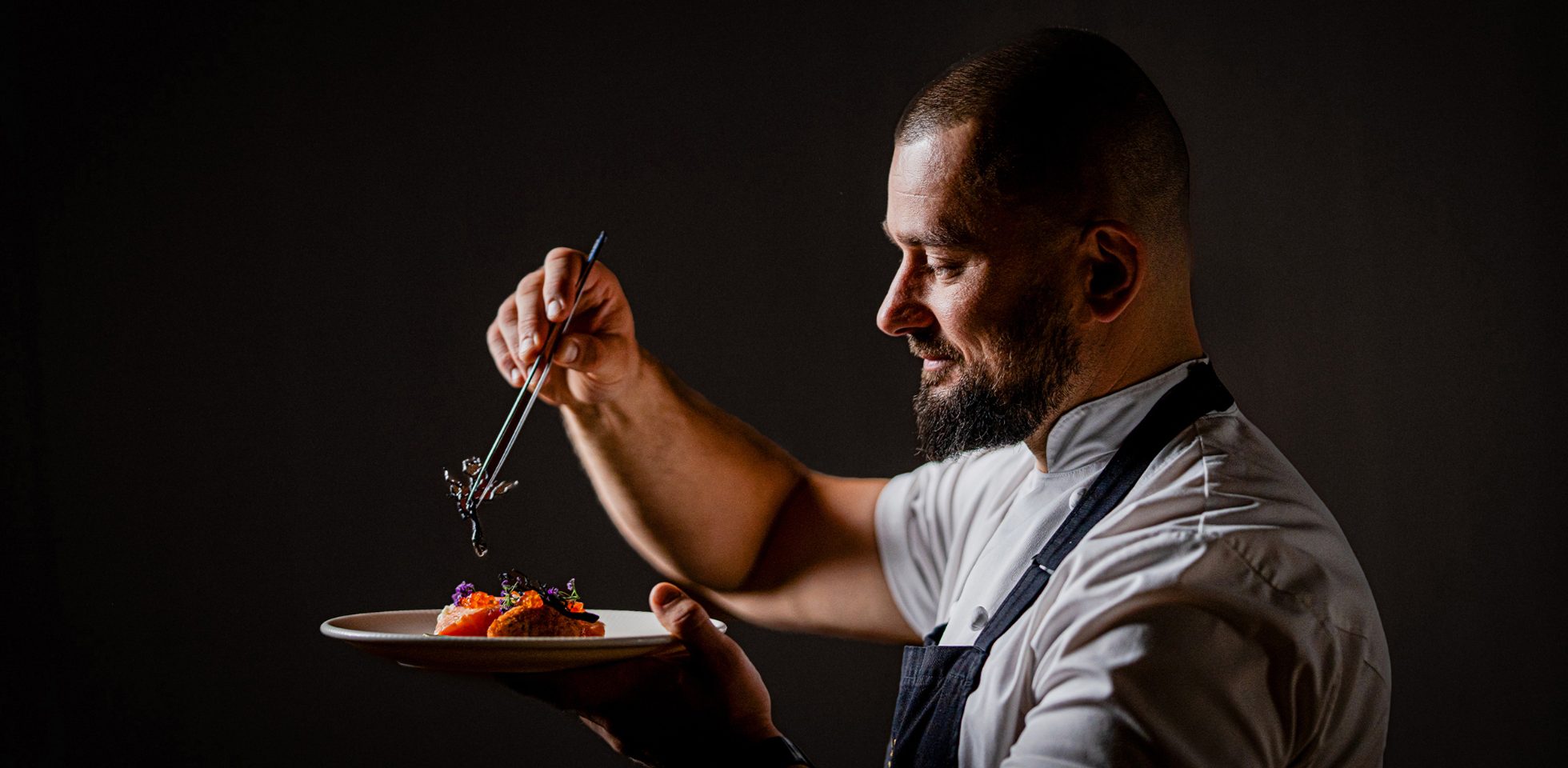 Szef Kuchni restauracji Rozbrat20 Bartosz Szymczak dekorujący swoje danie na ciemnym tle
