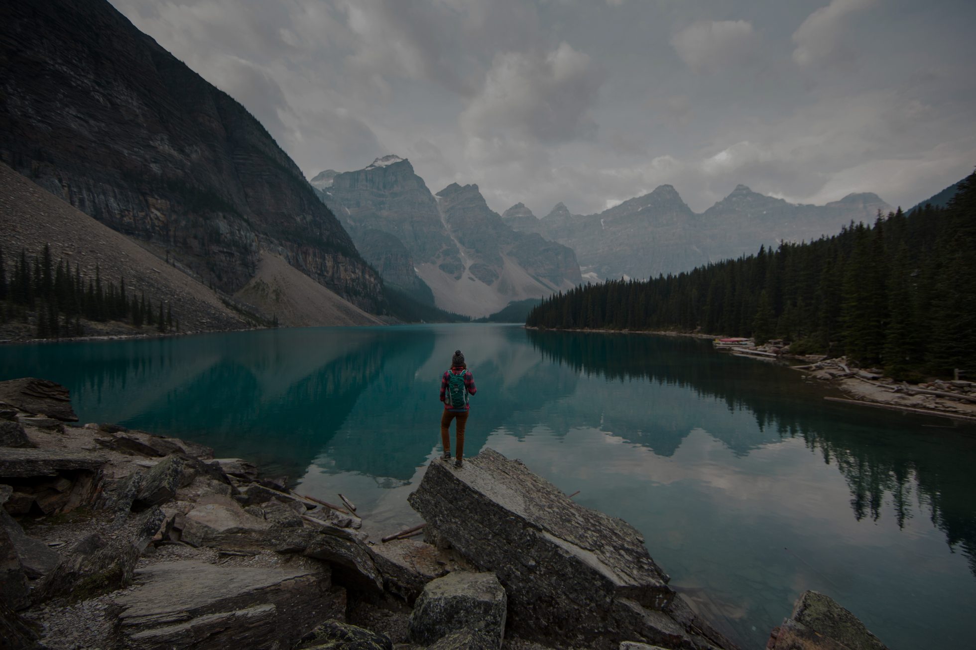 Una ragazza è in piedi in riva a un lago in Canada. Il lago è circondato da montagne. La donna è rivolta verso l'esterno ed è vestita con caldi abiti da trekking.